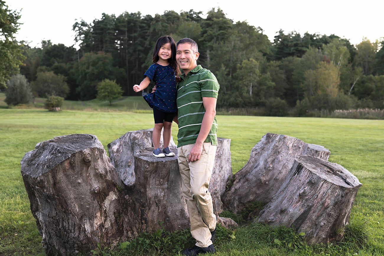 Little girl smiles on top of a tree stump as her and her dad pose for a photo