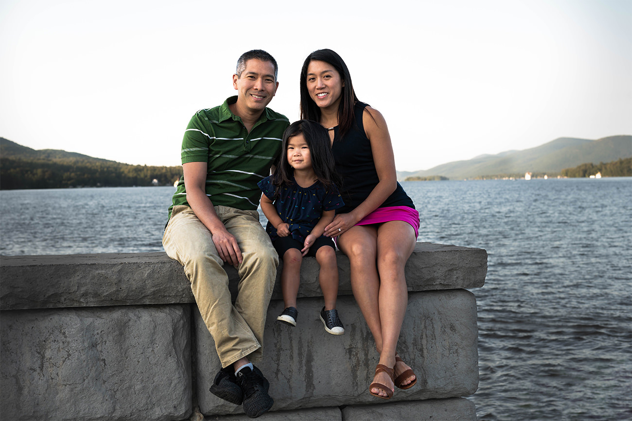 Family photography session in Lake George with husband, wife, and child posing in front of water.